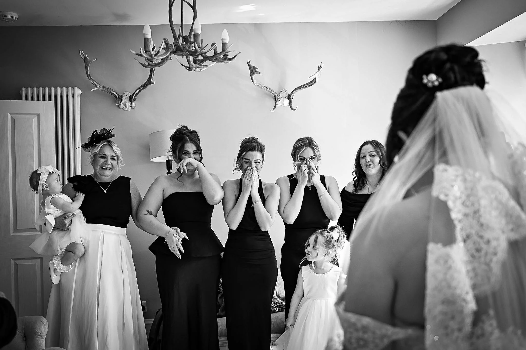 A bride in a lace dress is facing a group of five women, four of whom are visibly emotional, and two young girls, in a room with antler chandeliers on the wall.