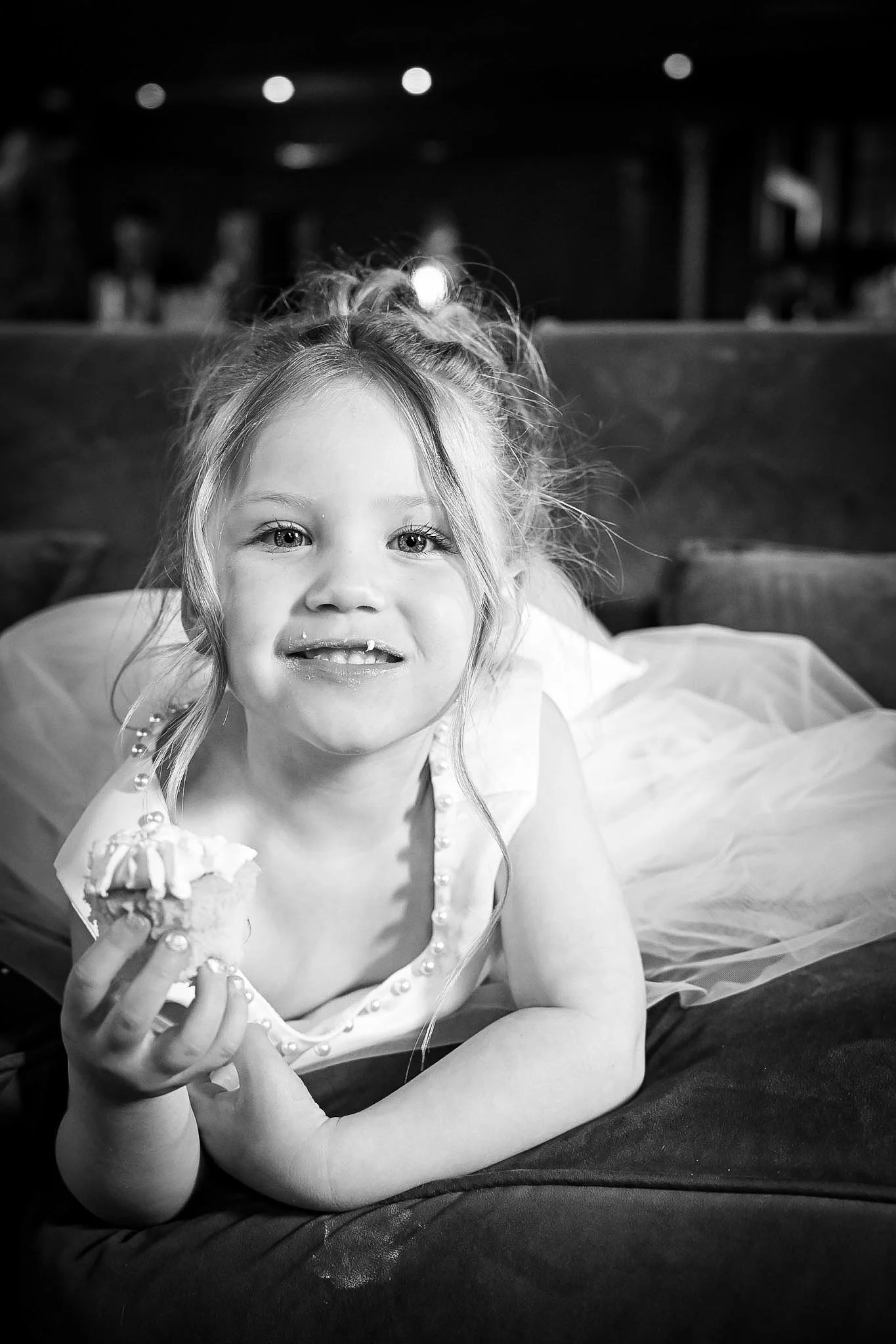 A young girl lying on her stomach, holding a cupcake, smiling with a gap-tooth smile, in a celebration or party setting, in black and white.