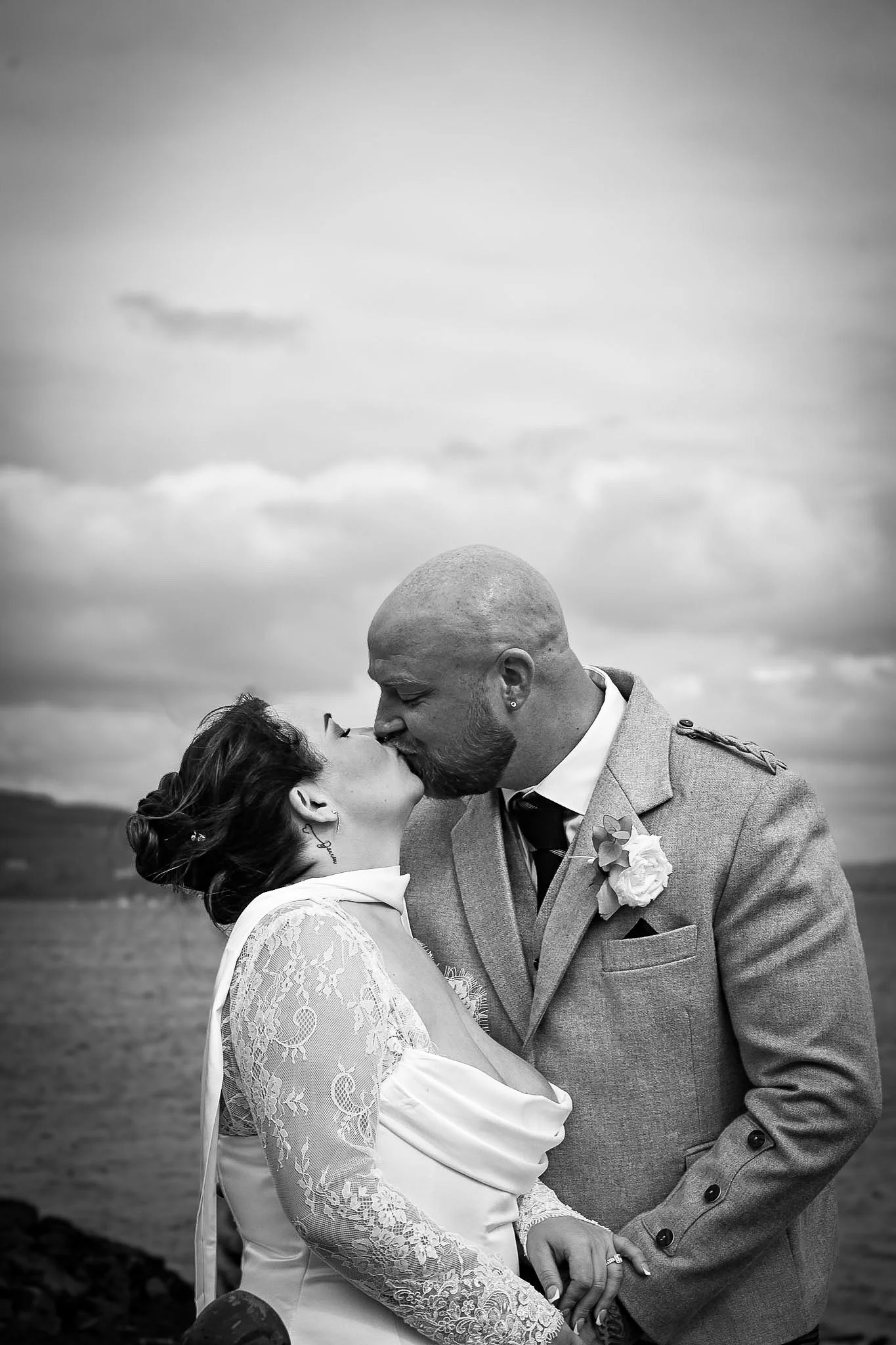 Black and white photo of a couple kissing outdoors, with water and cloudy sky in the background. The woman wears a lace dress and has her hair styled up, while the man wears a suit with a boutonniere.