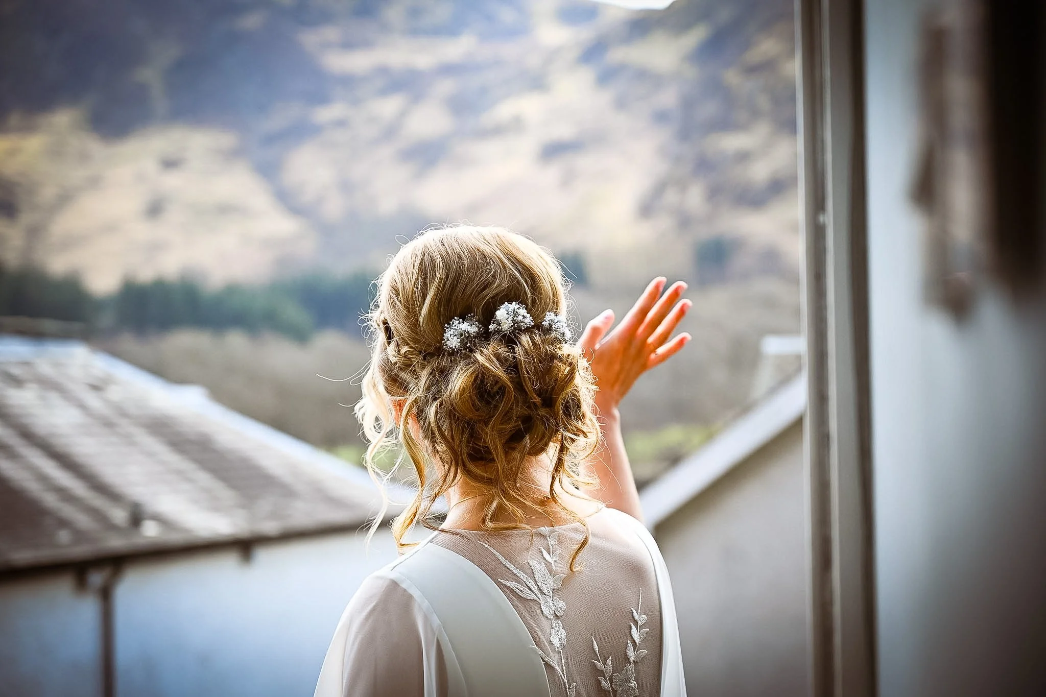 A woman with curly hair and floral hair accessories, wearing a white dress with lace details, looks out a window at a mountainous landscape, wiping away tears.