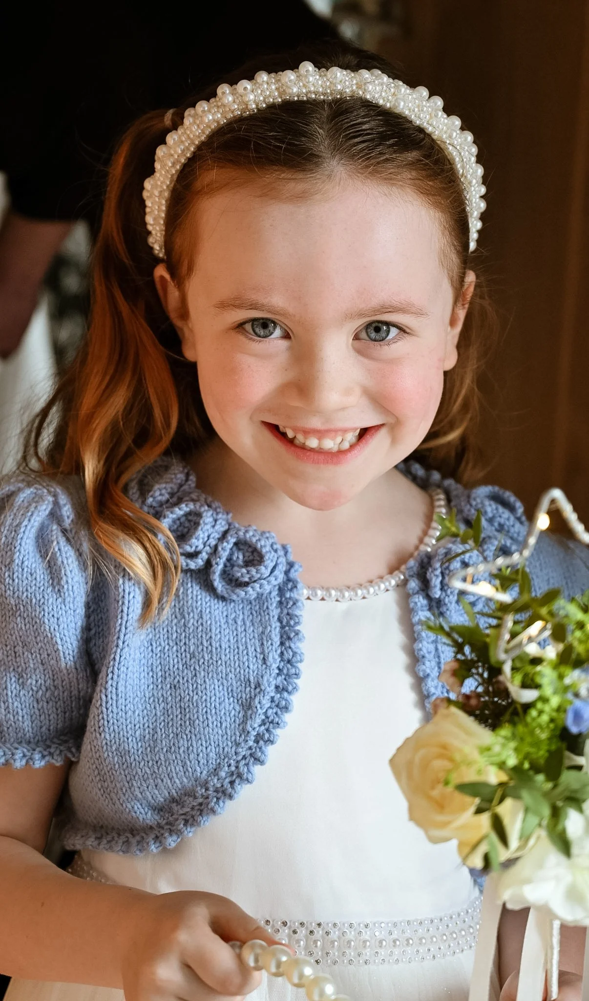 A young girl with red hair, blue eyes, and a big smile, wearing a pearl headband and a blue knitted cardigan over a white dress, holding a bouquet of flowers and a pearl necklace.