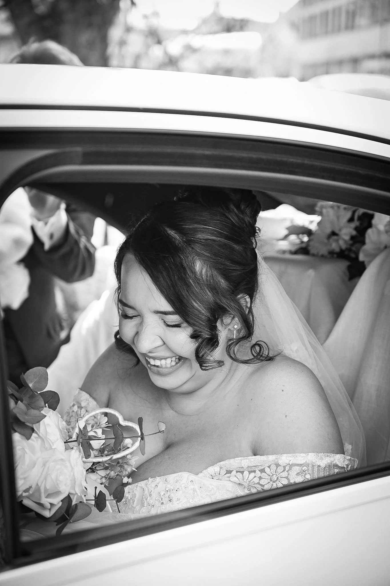 A bride sitting in a car, smiling and laughing, holding a bouquet of flowers.