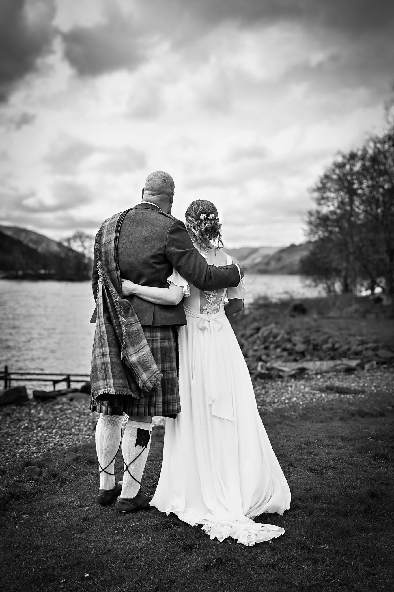 A black and white photo of a couple in wedding attire, standing by a lake with hills and trees in the background, embracing each other.