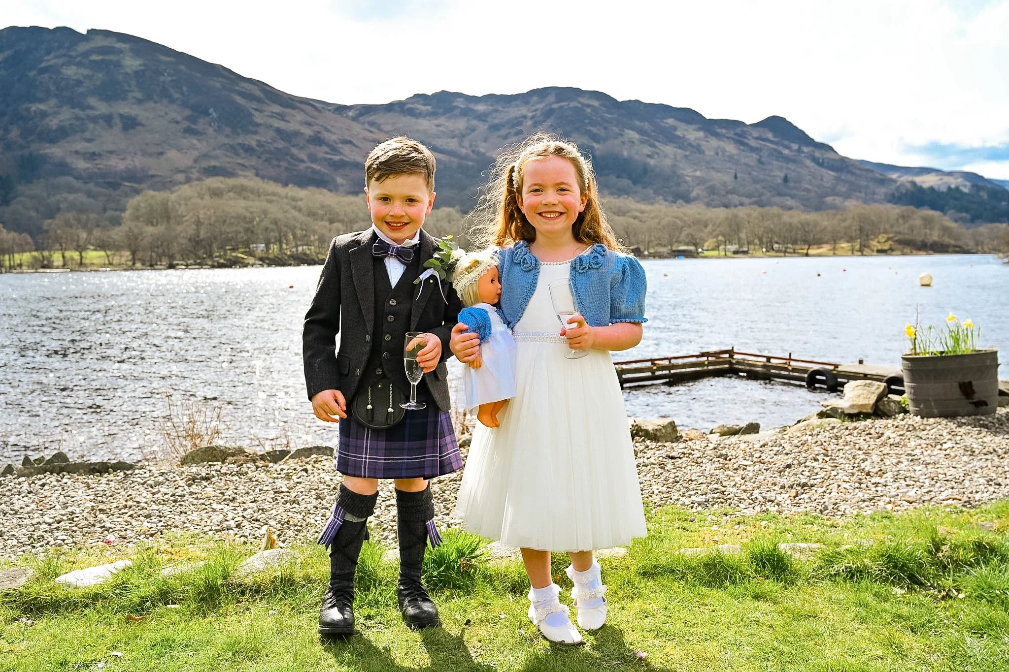 Two children, a boy and a girl, dressed in formal attire, smiling and holding drinks, standing on grass by a lake with mountains in the background