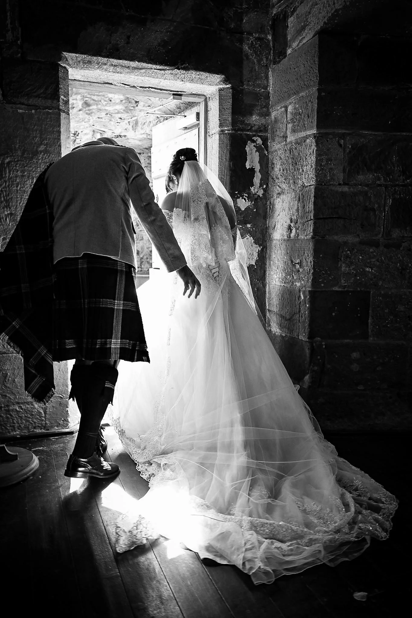 A bride in a wedding gown and veil standing in a doorway with a man, possibly her partner, adjusting her gown inside a stone building.