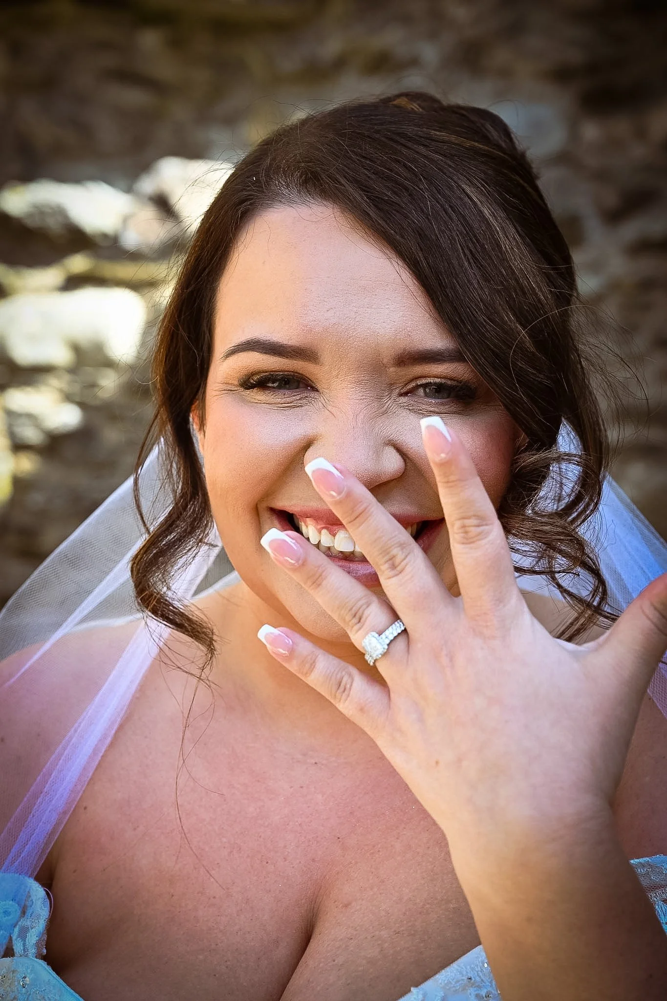 A woman smiling in a wedding dress showing her engagement ring. She has her hand near her face with nails painted French manicure. Her hair is styled in loose curls.