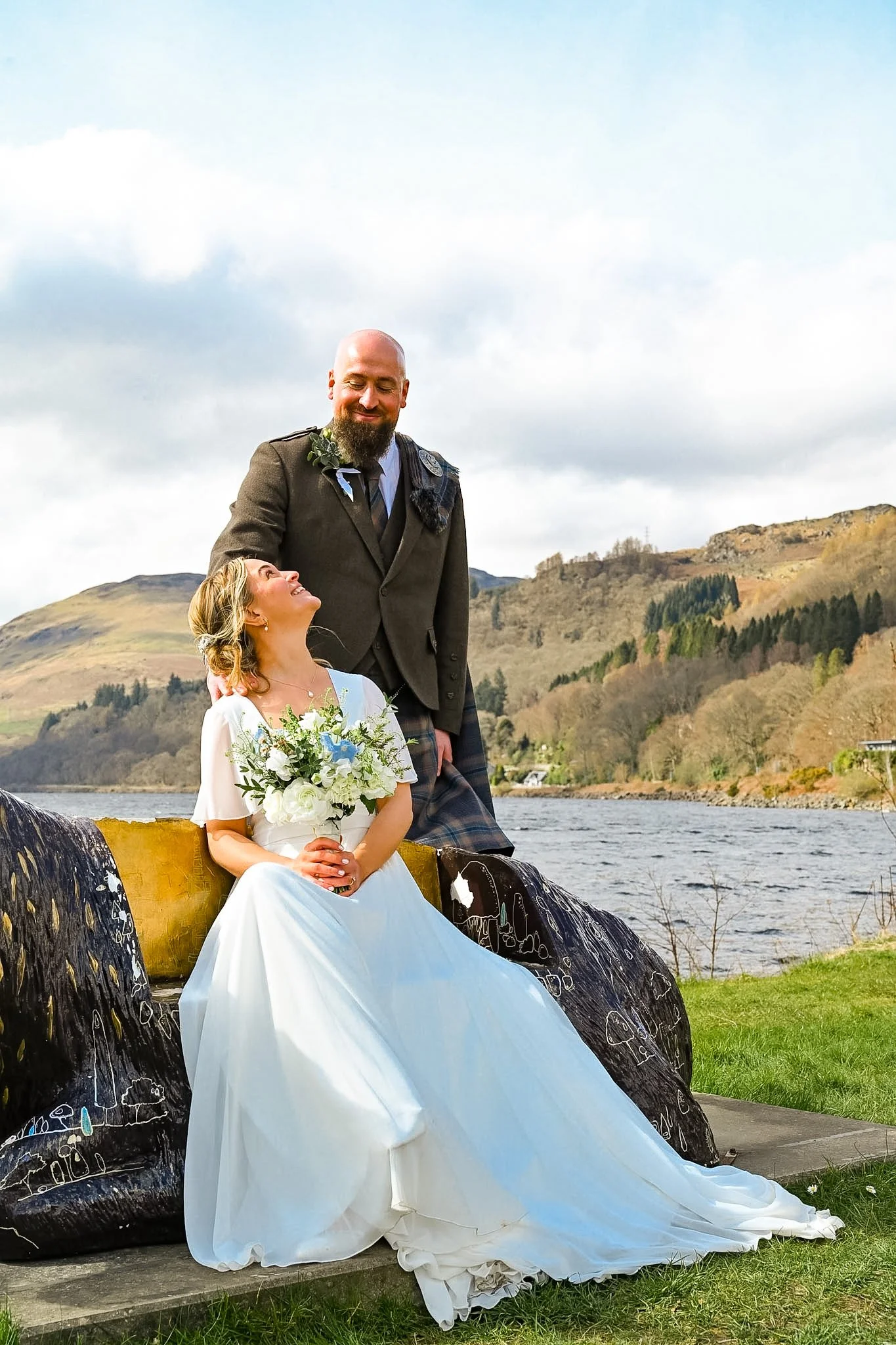 A bride sitting on a carved wooden bench holding a bouquet, smiling up at a groom standing beside her, outdoor setting with a river and hills in the background.