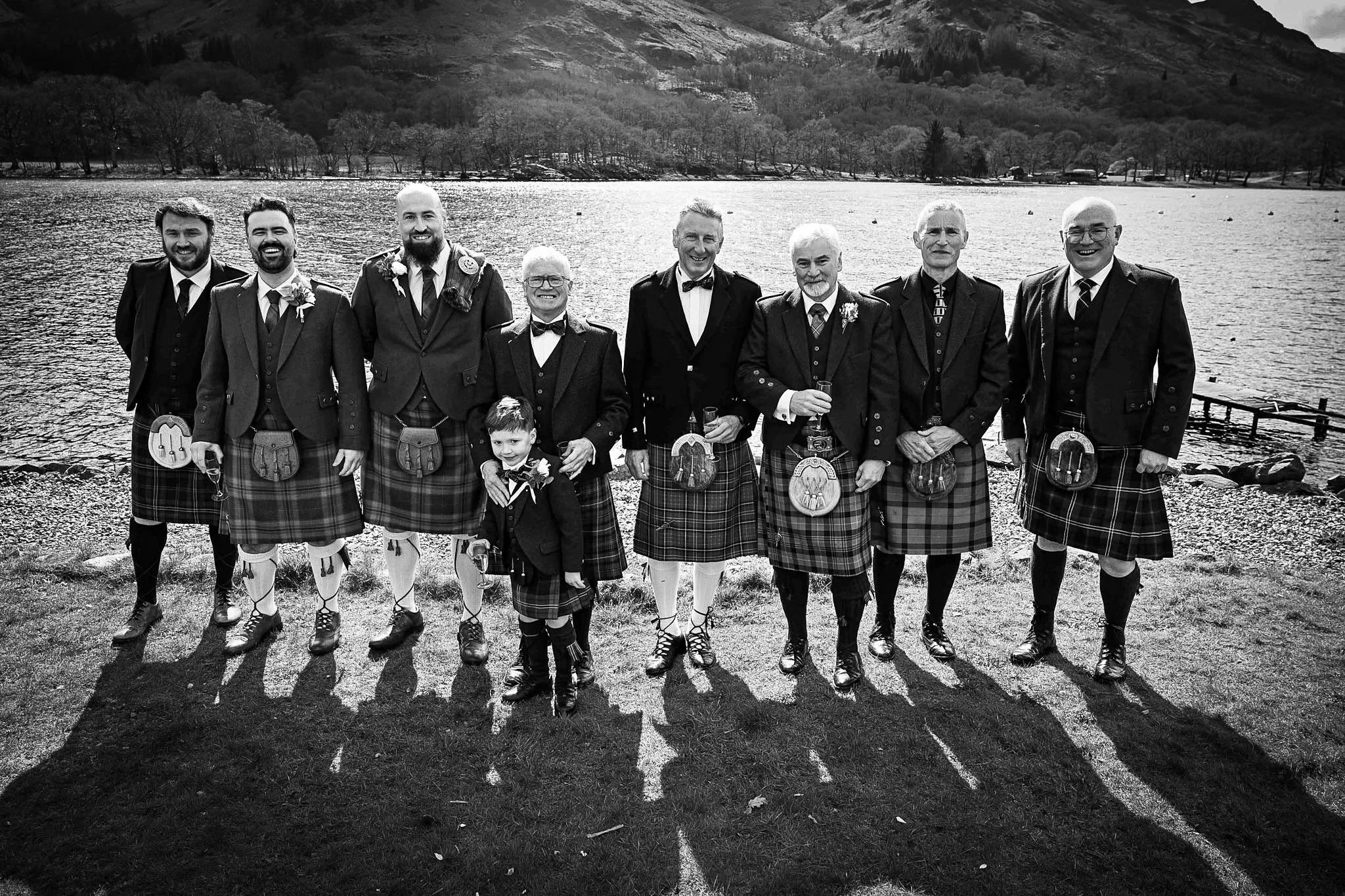 A group of men and two children dressed in traditional Scottish attire, including kilts, standing outdoors near a lake with hills and trees in the background on a sunny day.