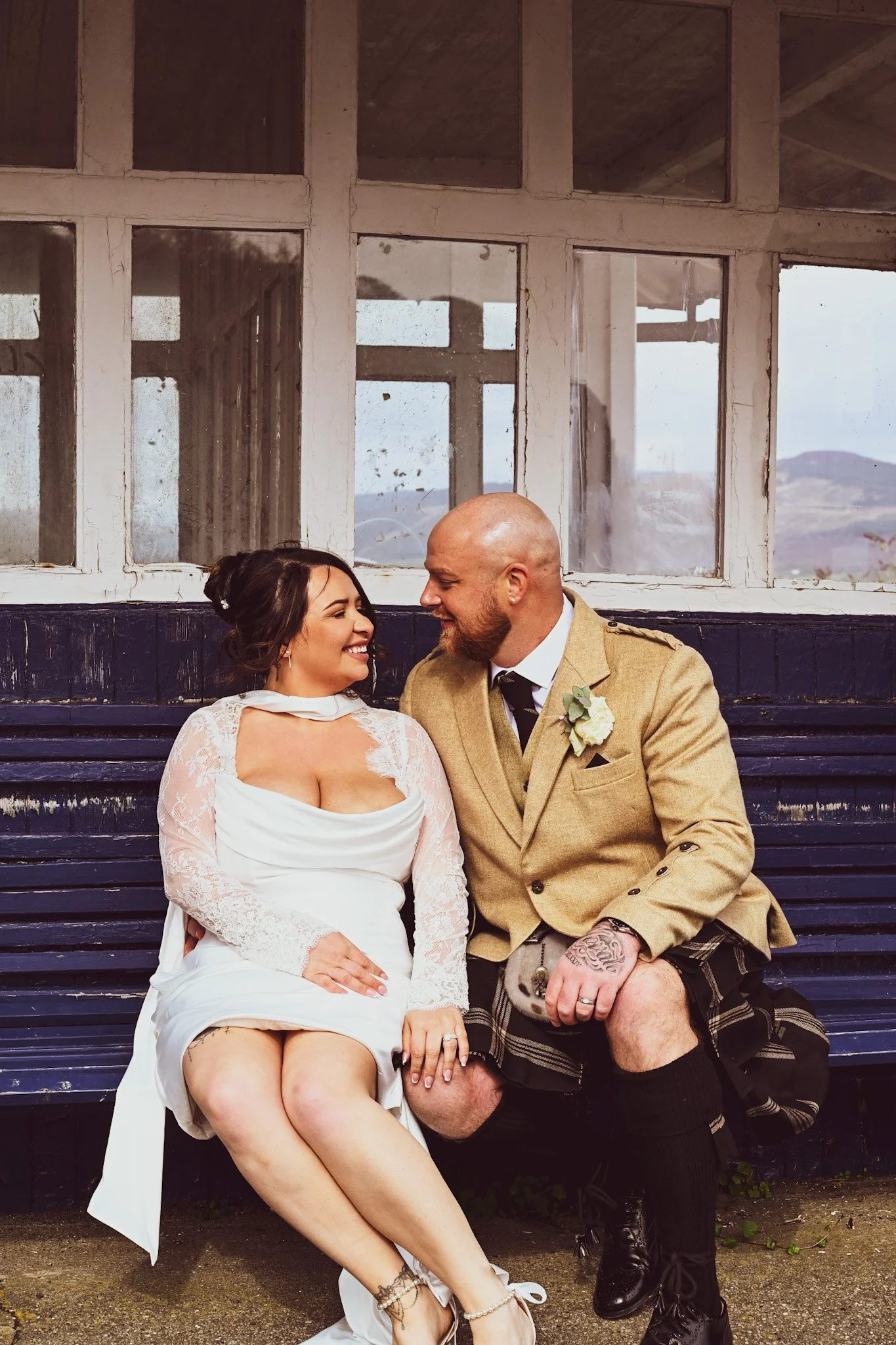 A bride and groom sitting on a wooden bench, smiling and looking at each other, with the bride in a white wedding dress and the groom in traditional Scottish attire, outside with a building and mountains in the background.