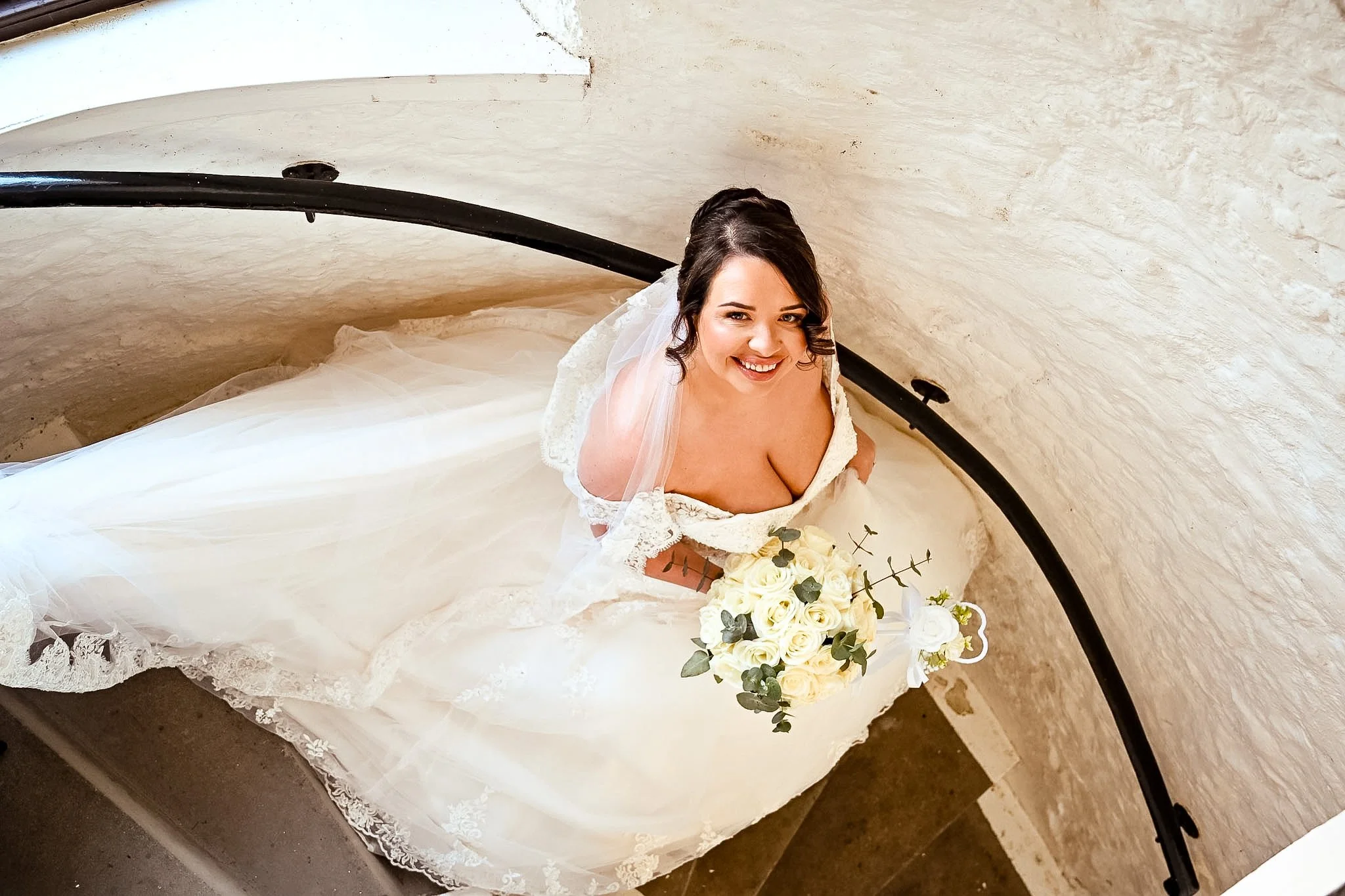 A smiling bride with dark hair in an updo, wearing a white lace wedding gown with a sweetheart neckline, holding a bouquet of white roses and greenery, standing on a staircase with a curved black railing and textured beige walls.