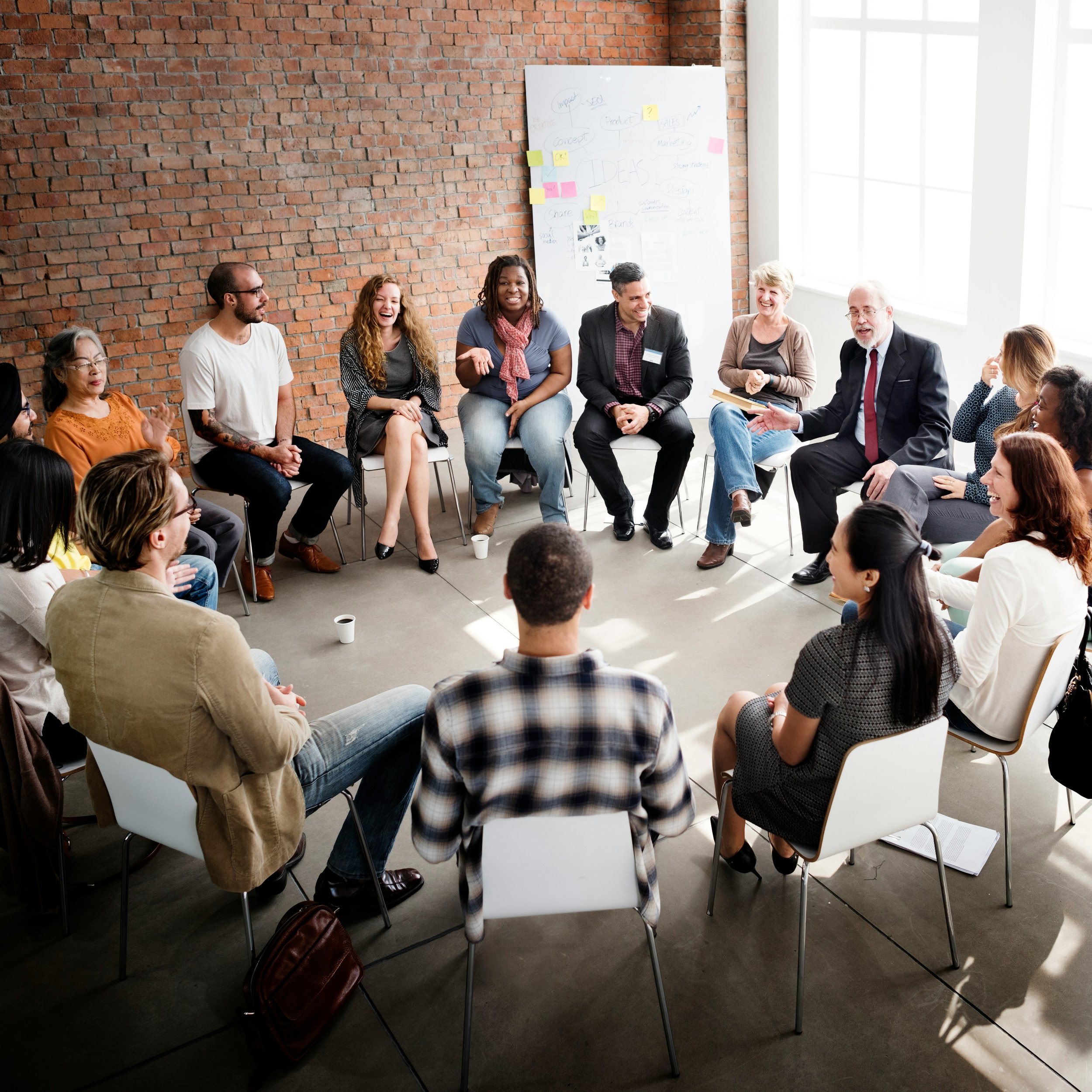 Group of diverse people sitting in a circle in a bright room with an exposed brick wall and a large window, engaging in a discussion or meeting.