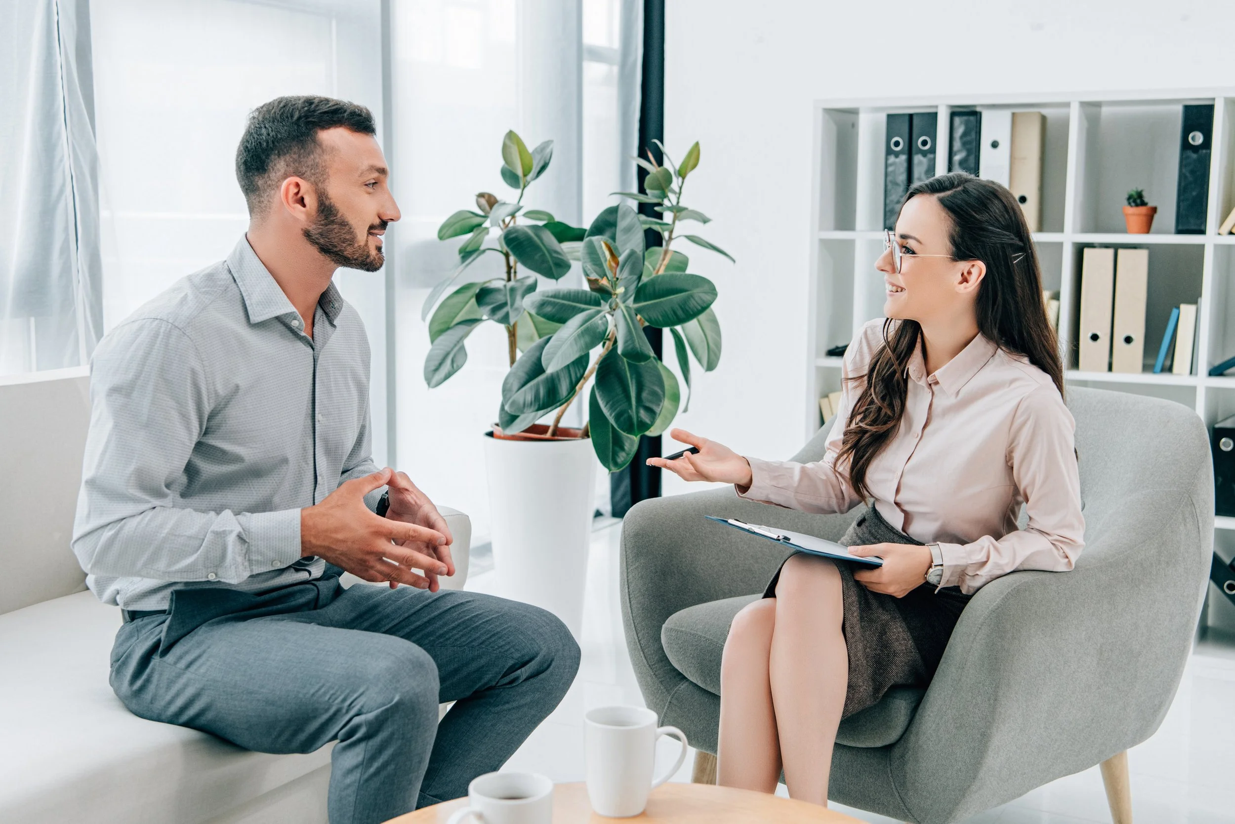 A man and woman having a conversation in a bright, modern office space. The woman is holding a clipboard and gesturing with her hand while the man is listening attentively.