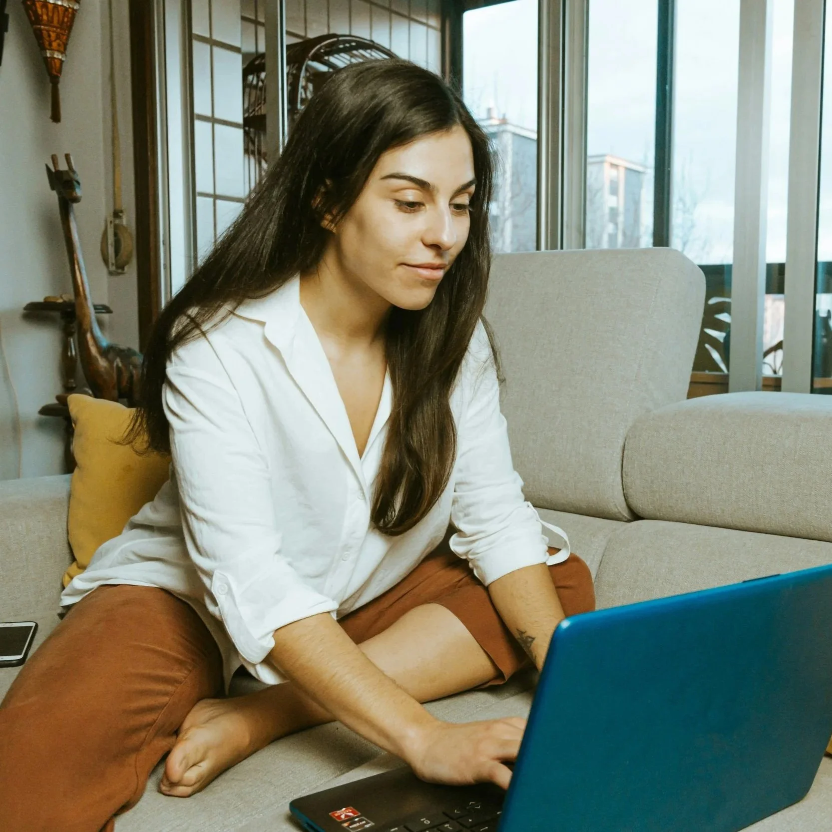 A young woman with long dark hair sitting on a beige couch, working on a blue laptop. She is wearing a white button-up shirt and brown pants. The room has large windows, with a wooden decor and a giraffe sculpture behind her.