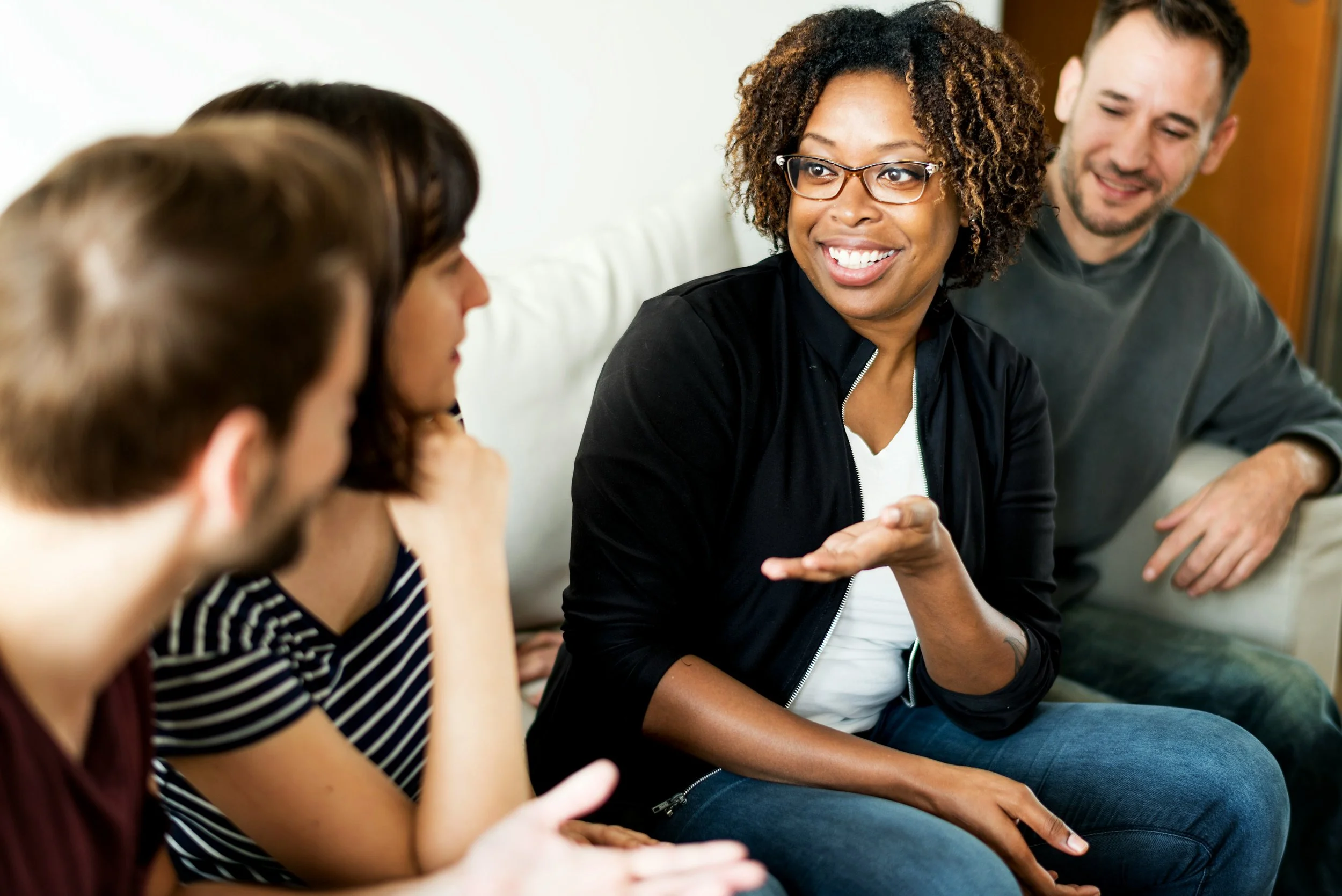 Group of four people sitting and engaging in conversation, woman with glasses smiling, others listening, casual setting.