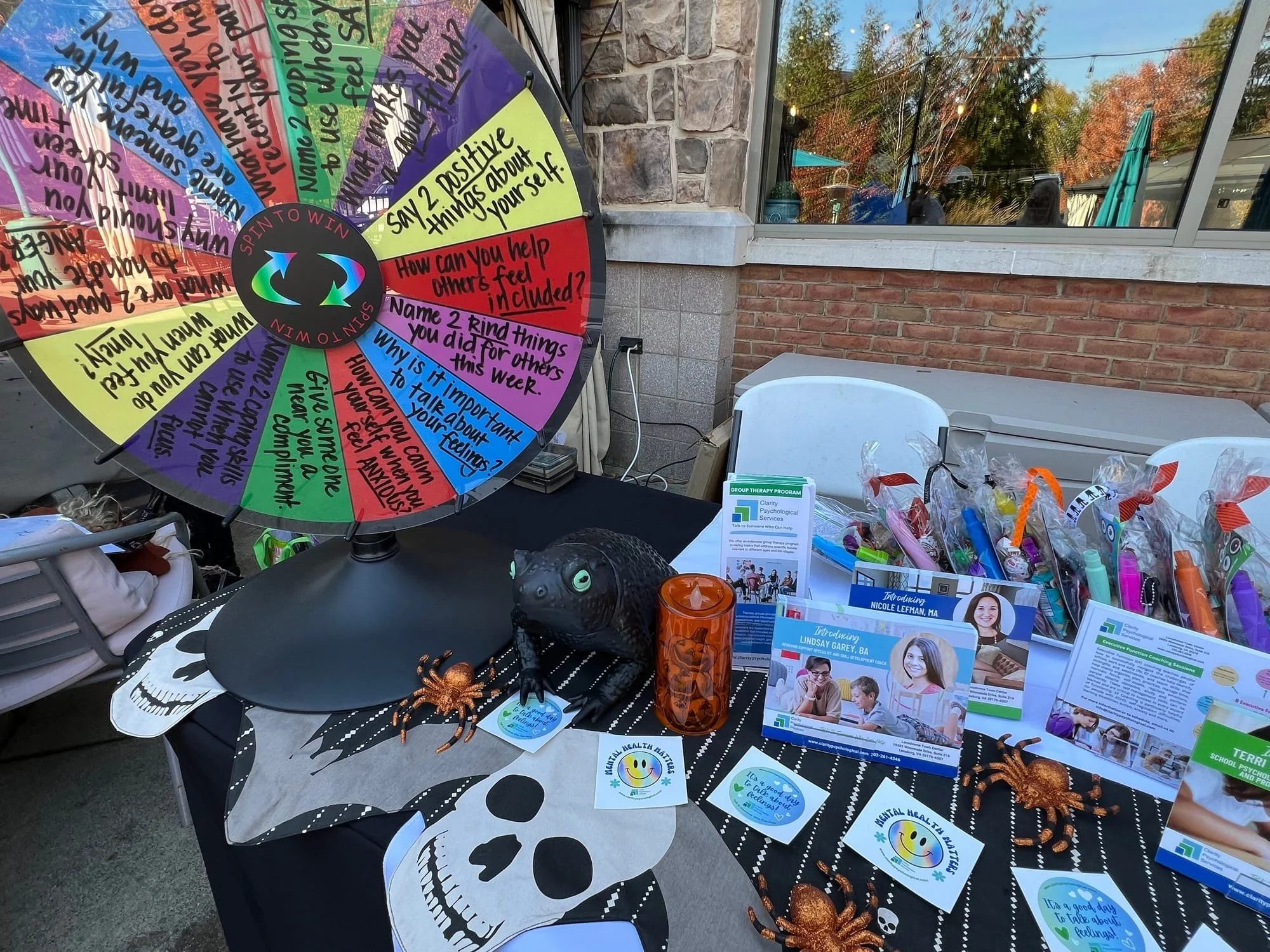 A table at an outdoor event displaying promotional materials about mental health awareness, including a colorful spinning wheel with questions and prompts, Halloween decorations like plastic spiders and a skull, and brochures with smiling people and contact information. The background shows a brick building with a large window and trees with autumn foliage.