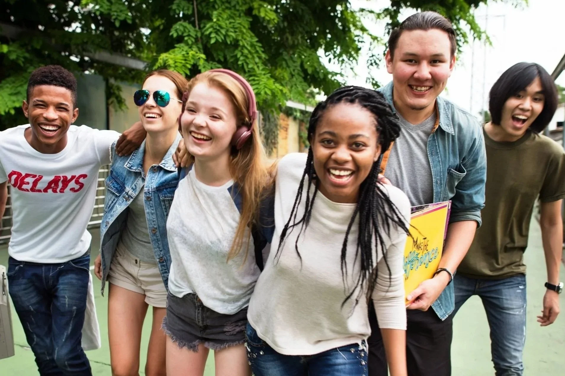 A diverse group of six young people walking outdoors with smiles and laughter, surrounded by greenery and trees.