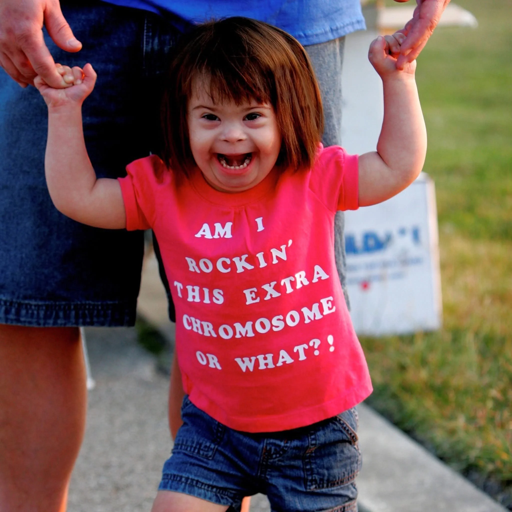 Close-up of a young girl with red hair wearing a pink t-shirt and denim shorts, holding hands with an adult, outdoors during a sunny day.