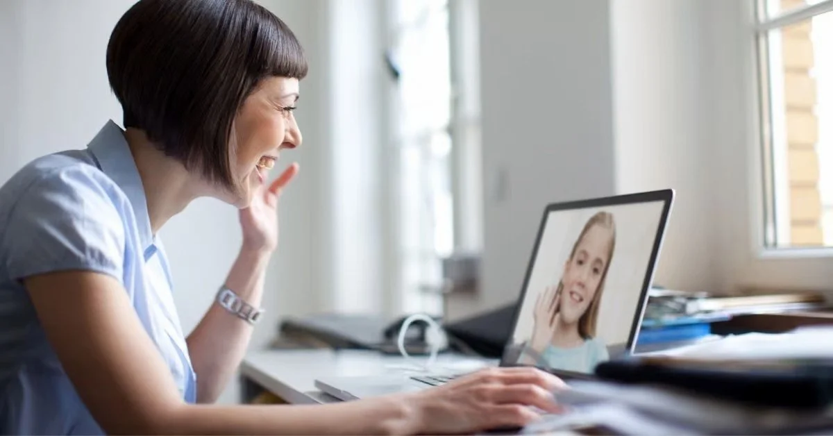 A woman with short brown hair smiling during a video call on her laptop, which shows a young girl with long blonde hair on the screen.
