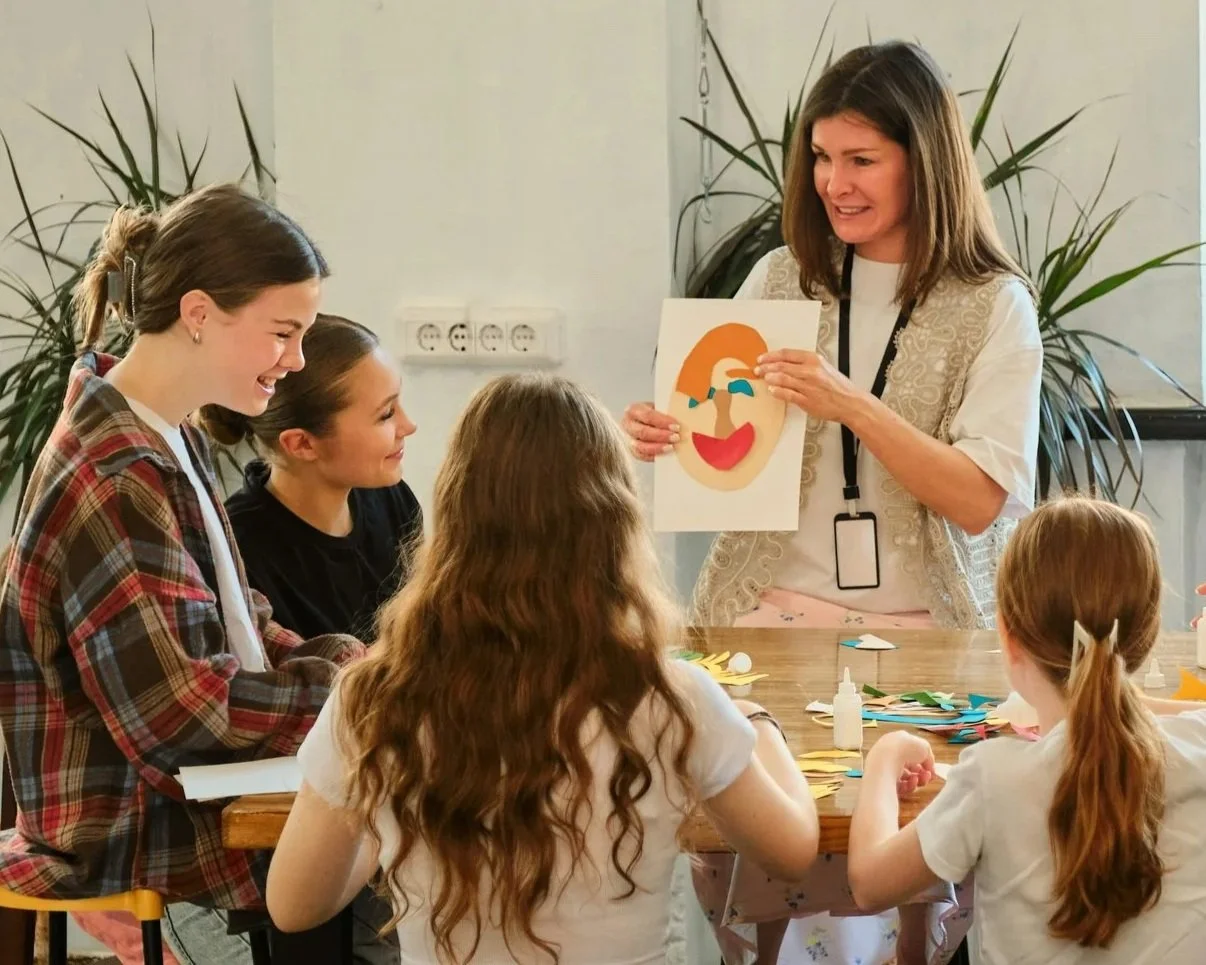 A woman shows children paper crafts of a smiling face with tears and a nose. The children sit at a table covered with paper and craft supplies, smiling and engaged in an art activity.