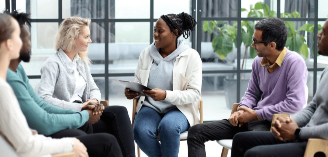 Group therapy session with six diverse people sitting in a circle, a woman in the center holding a tablet and smiling.