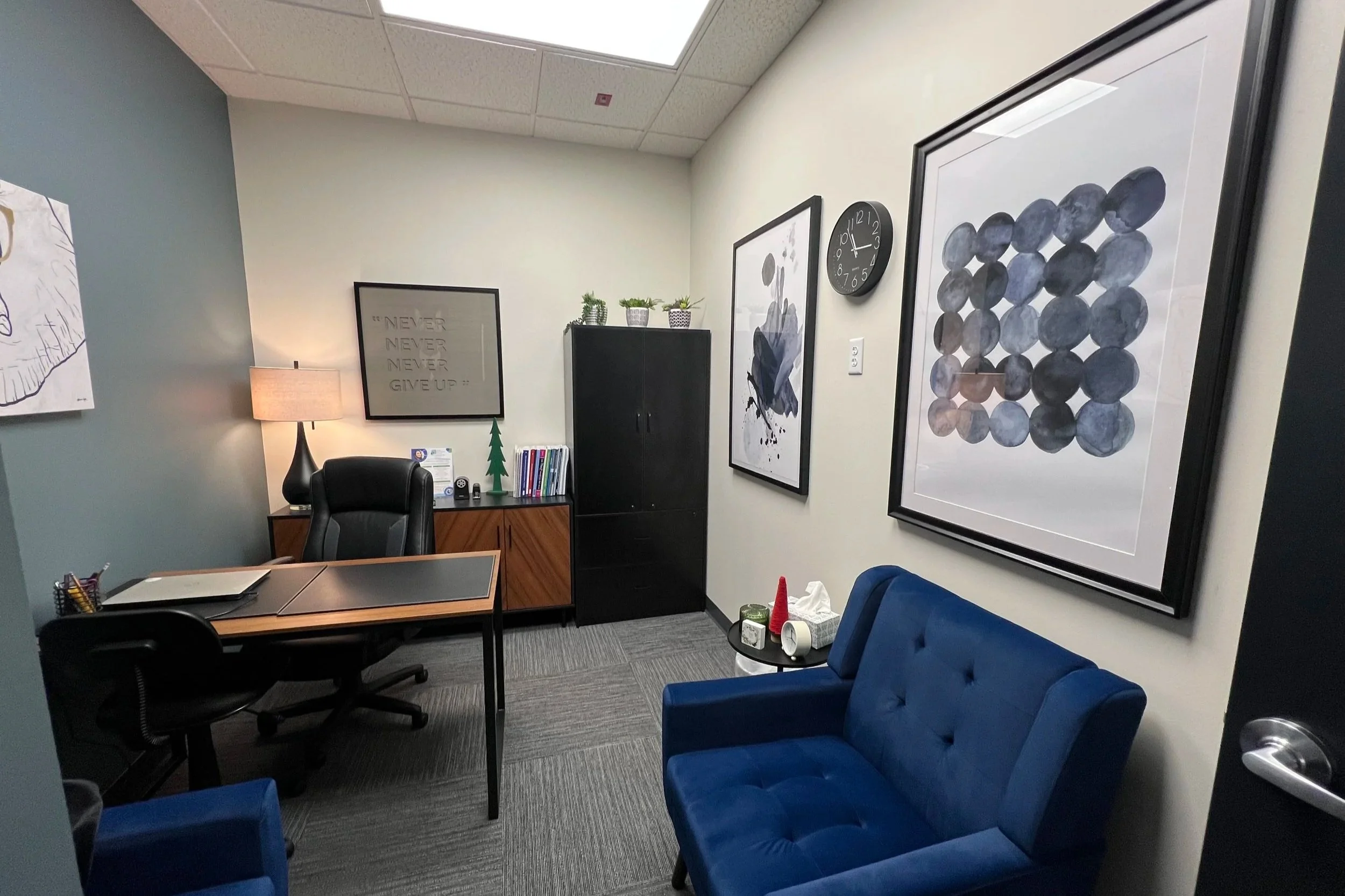 An office room with a desk, a black office chair, and a blue couch. The wall has framed abstract art, a large clock, and some potted plants. A small table next to the couch holds tissues and decorative cones.