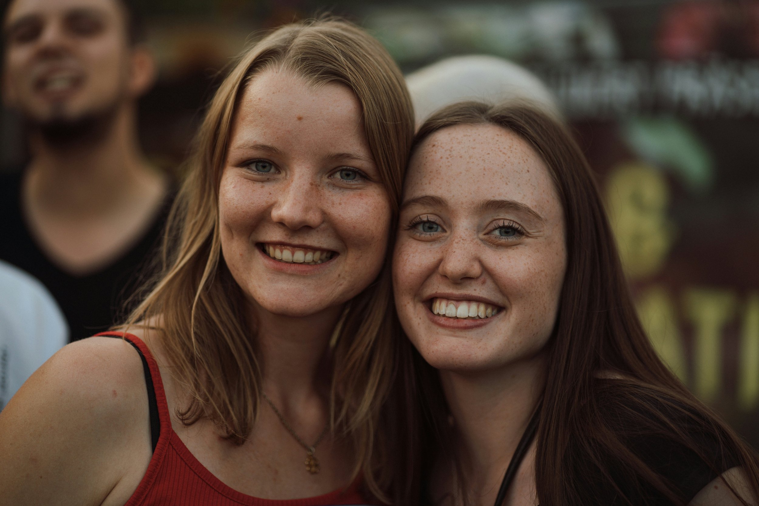 Close-up of two smiling young women with freckles, one with red hair and one with brown hair, taking a selfie together outdoors with a blurred background.
