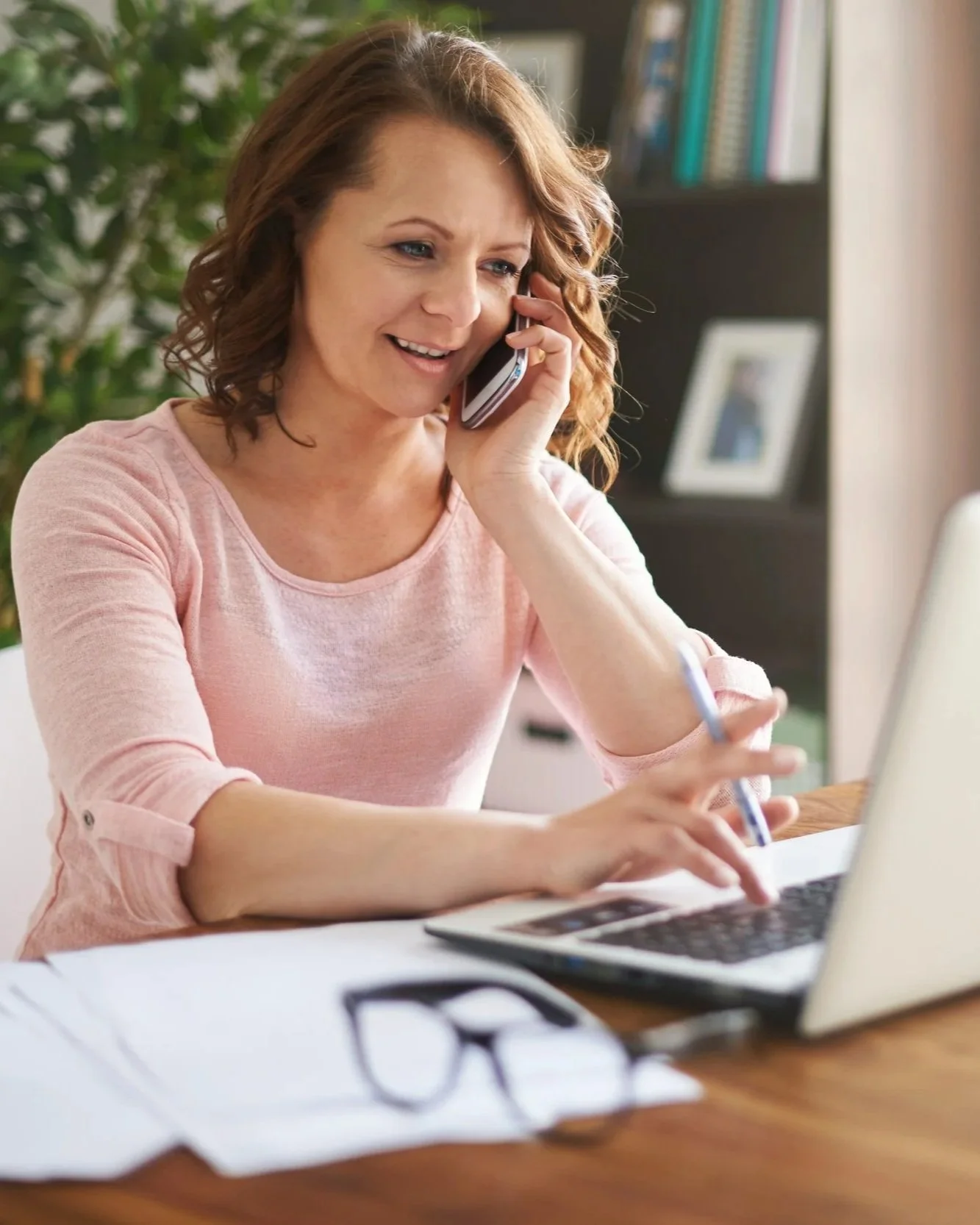 A woman with curly red hair, wearing a pink shirt, is talking on the phone while working on a laptop at a wooden desk in a well-lit room with a bookshelf and a large green plant in the background.