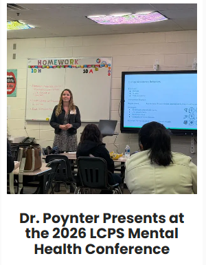 A woman presenting at a conference in front of a classroom with students seated at desks. There is a large screen displaying presentation slides and a whiteboard behind her with colorful writing and decorations.