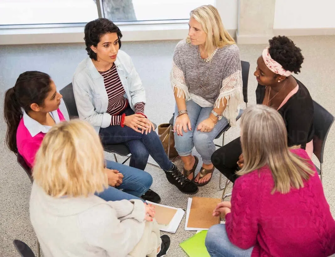 Six women sitting in a circle in a room having a discussion.