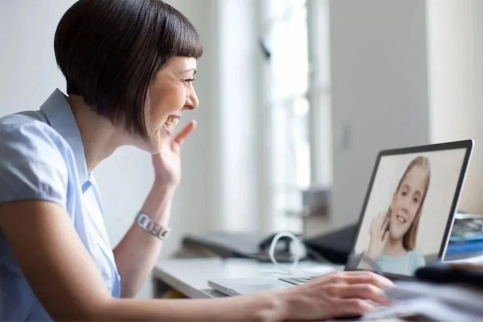 A woman with dark hair, wearing a light blue shirt, talking and smiling during a video call on her laptop with a young girl on the screen.