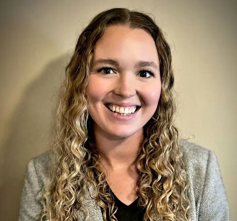 A woman with long curly blonde hair, smiling, wearing a gray blazer over a black top, standing in front of a beige wall.