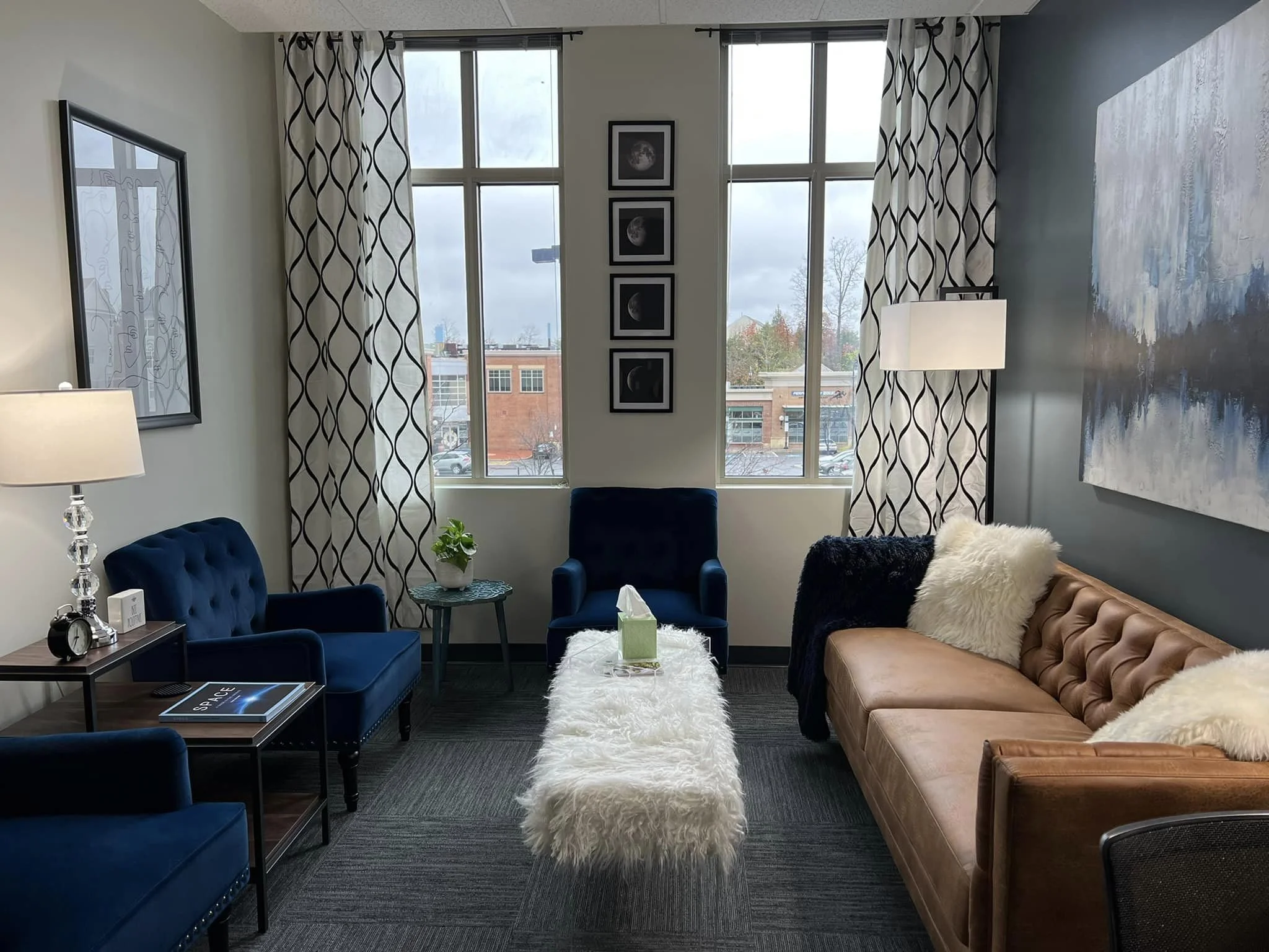 A cozy living room with large windows covered by white curtains with black geometric patterns. There are two blue velvet armchairs, a tan leather sofa with white and black furry pillows, a black side table with a lamp and clock, and a small white fluffy ottoman in the center. The walls are decorated with framed artwork and a large abstract painting, and there is a small plant on a shelf near the window.