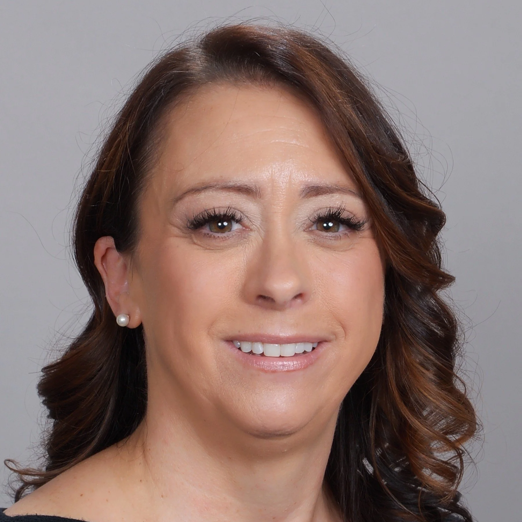 Nicole Lefman, BS - Headshot - A smiling woman with brown hair styled in loose curls, wearing pearl earrings, in front of a grey background.