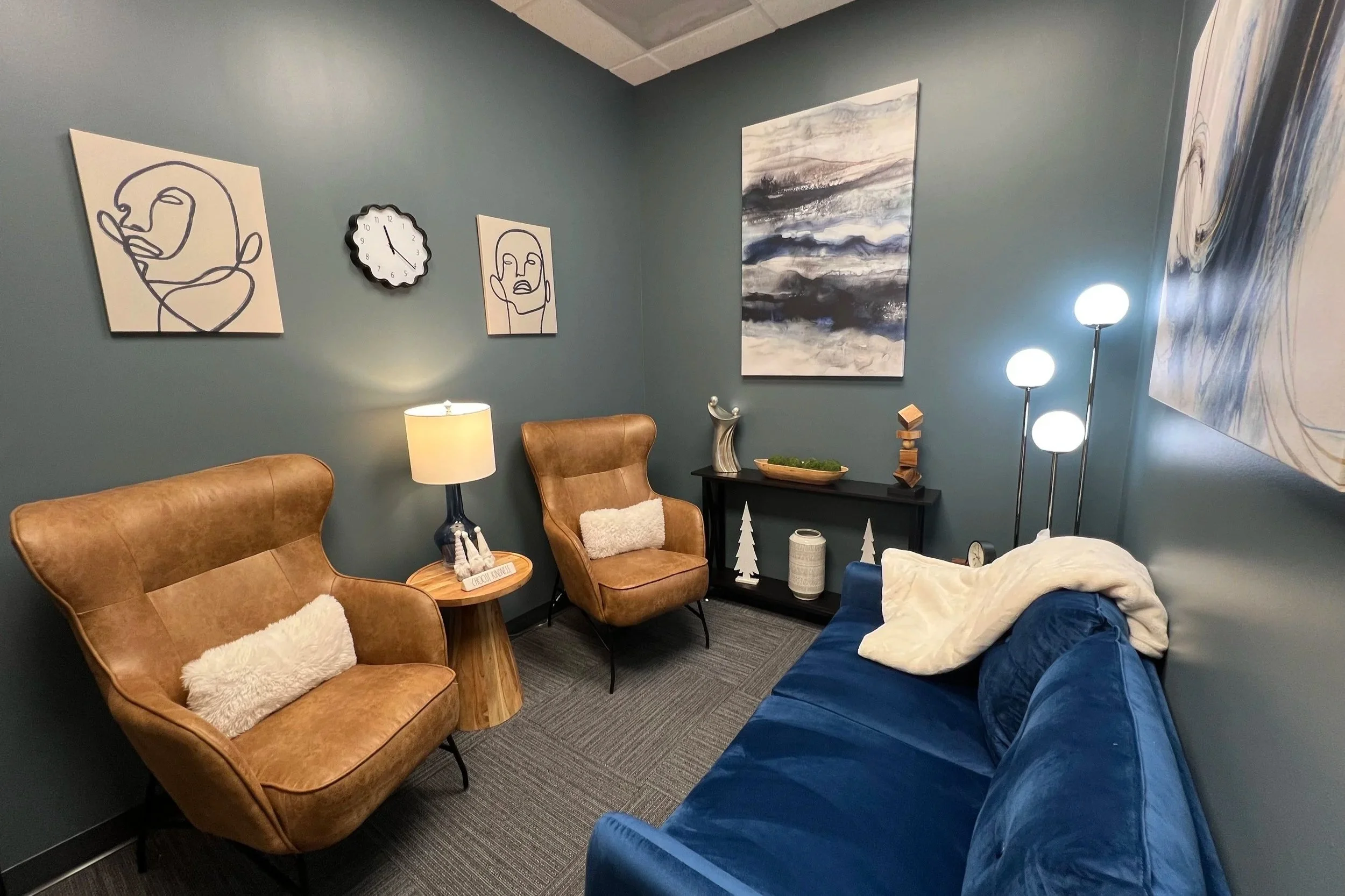A cozy living room corner with blue walls, featuring two tan leather armchairs with white fluffy pillows, a blue velvet sofa, a small round wooden side table with a lamp, and a black console table with decorative items. The wall is decorated with abstract art and a clock showing 12:02, plus a tall floor lamp with three white globe lights.
