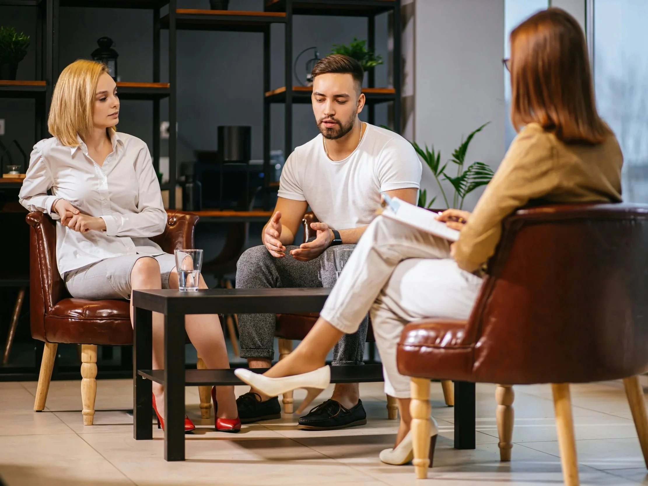 Three people having a discussion in a modern indoor setting, with two women and one man sitting on chairs around a small table. The woman on the left has blonde hair and is wearing a white shirt, the man in the middle has dark hair and a beard, wearing a white t-shirt, and the woman on the right has reddish-brown hair and is wearing glasses and a yellow top. The background includes decorative shelving with plants and a large window.