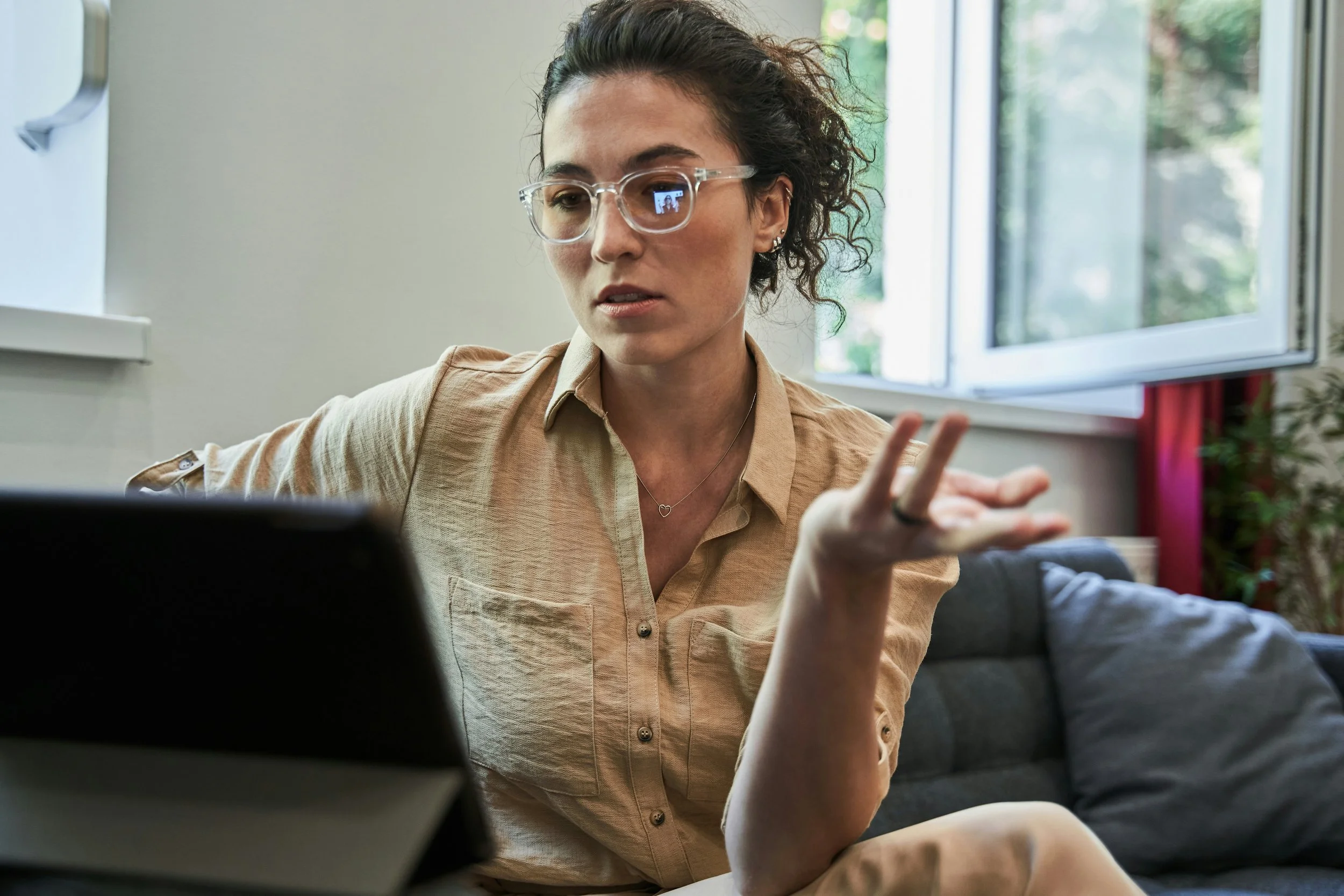 A woman with curly hair and glasses working on a laptop in a room with open windows and natural light.
