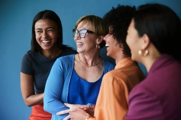 Four women smiling and chatting together indoors against a blue wall.