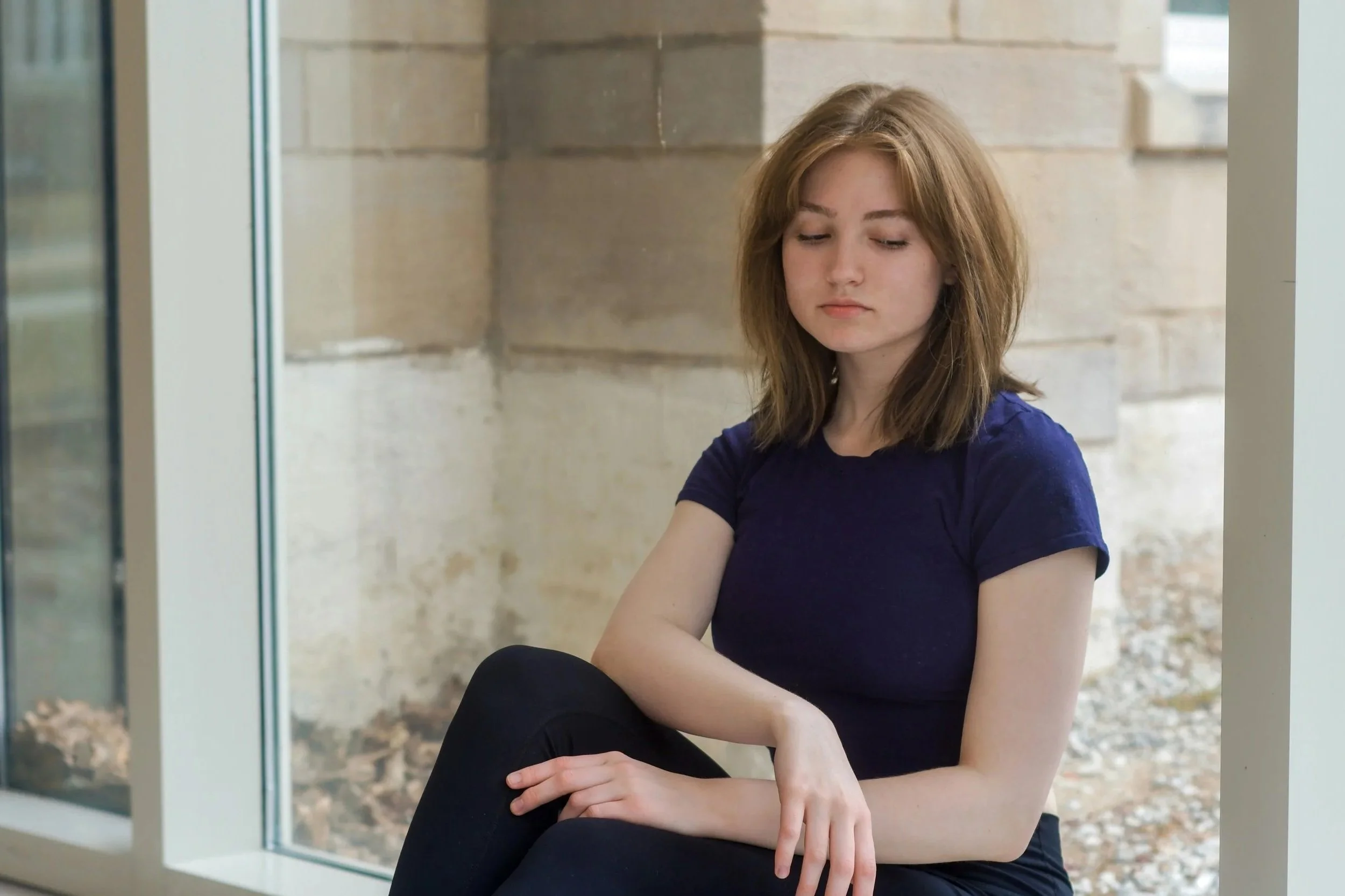 A young woman with shoulder-length reddish-brown hair, wearing a dark blue t-shirt, sitting by a large window with a stone wall behind her, looking down with a thoughtful expression.