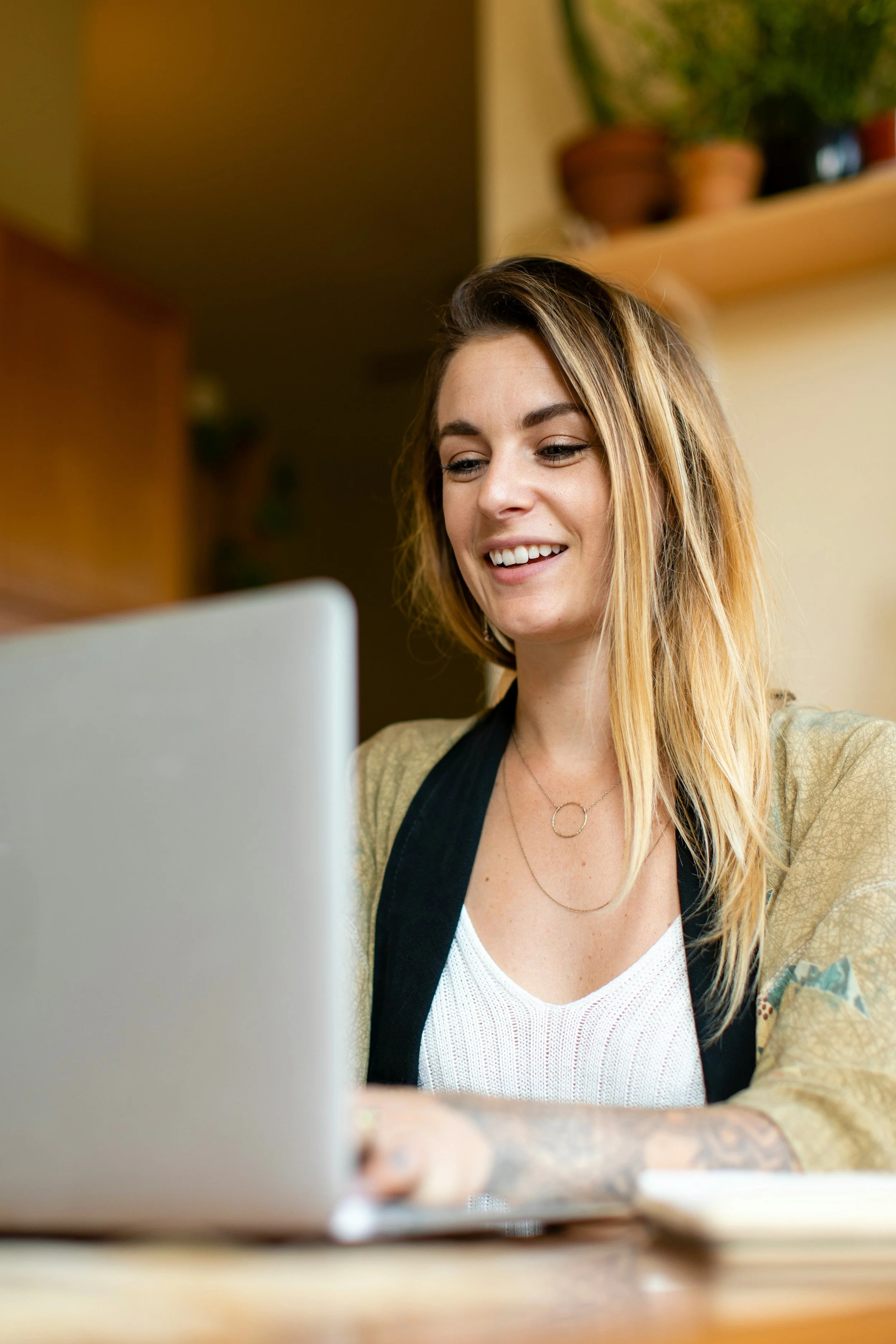 A young woman with long blonde hair smiling while working on a laptop at a desk in a cozy, well-lit room. Potted plants are visible on a shelf in the background.
