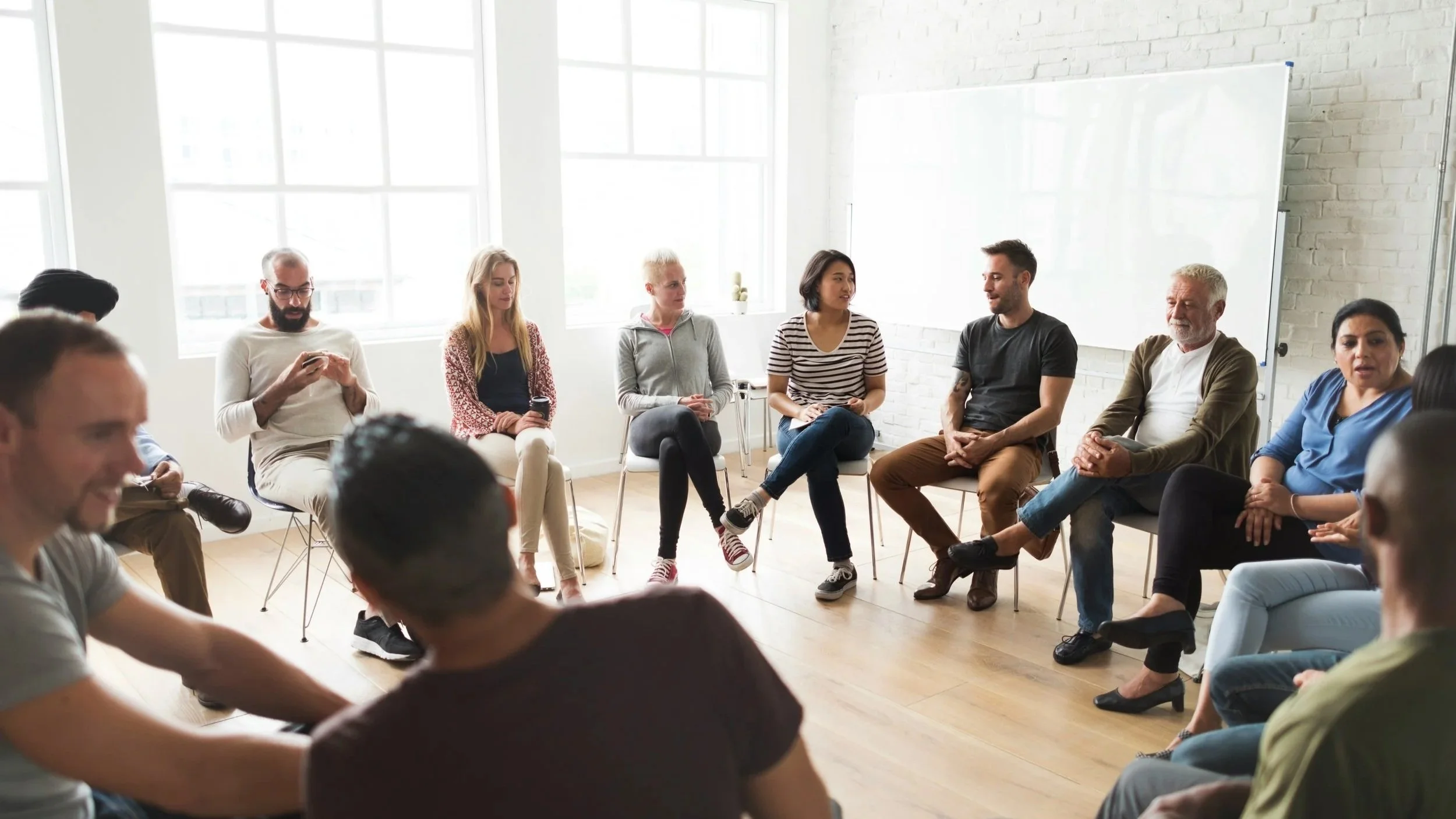 A diverse group of people sitting in a circle in a bright, spacious room with large windows and white brick walls, engaging in a discussion or meeting.
