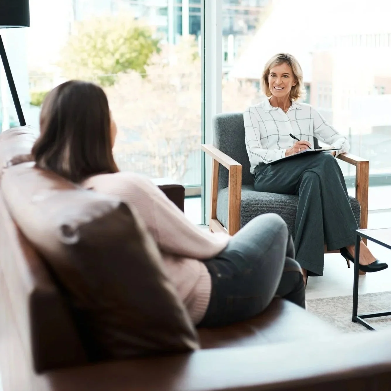 A professional woman conducting a counseling or interview session with a client in a modern office setting with large windows and a city view.