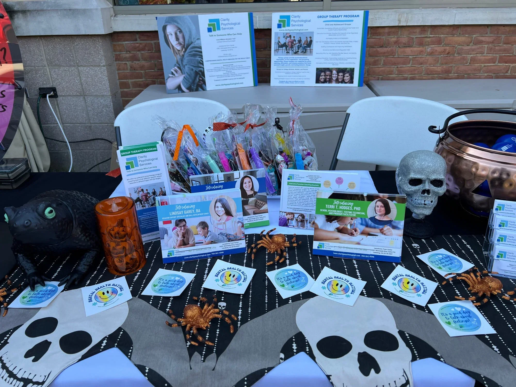 Table display promoting mental health and psychological services with flyers, business cards, and Halloween decorations including skull and spider figures, set against a brick wall and window.