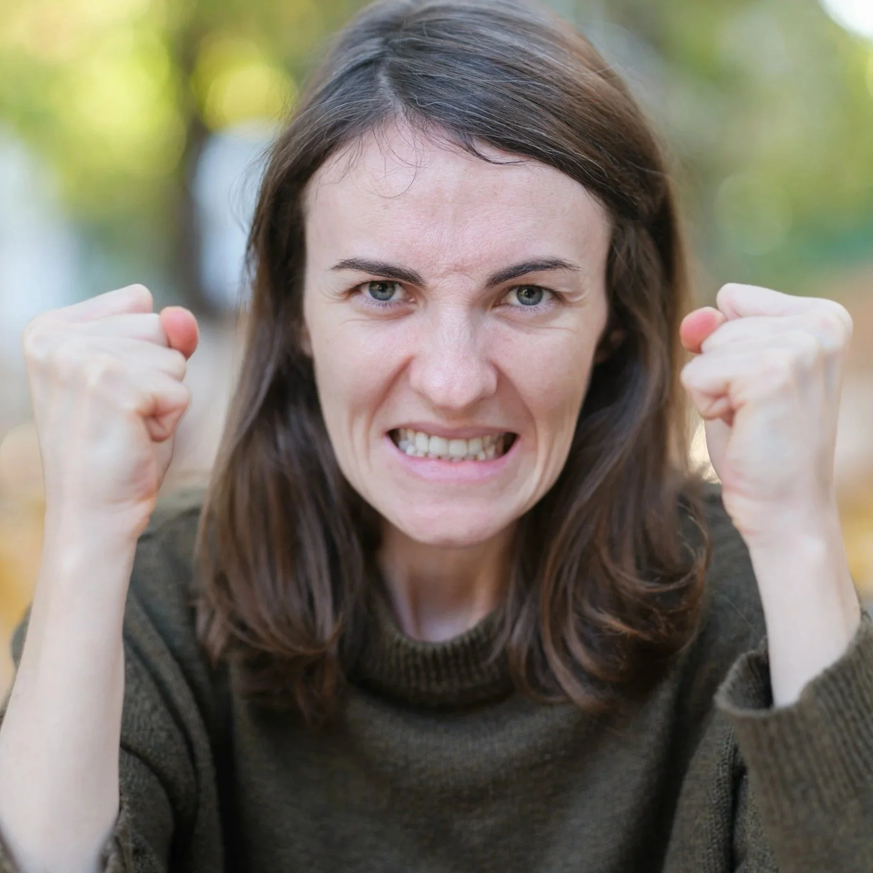 A woman with brown hair and blue eyes making a fierce facial expression and flexing her arm muscles outdoors with blurred green background.