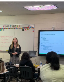 A teacher stands in front of a whiteboard in a classroom, delivering a lesson to students seated at desks, with a large screen displaying presentation slides.