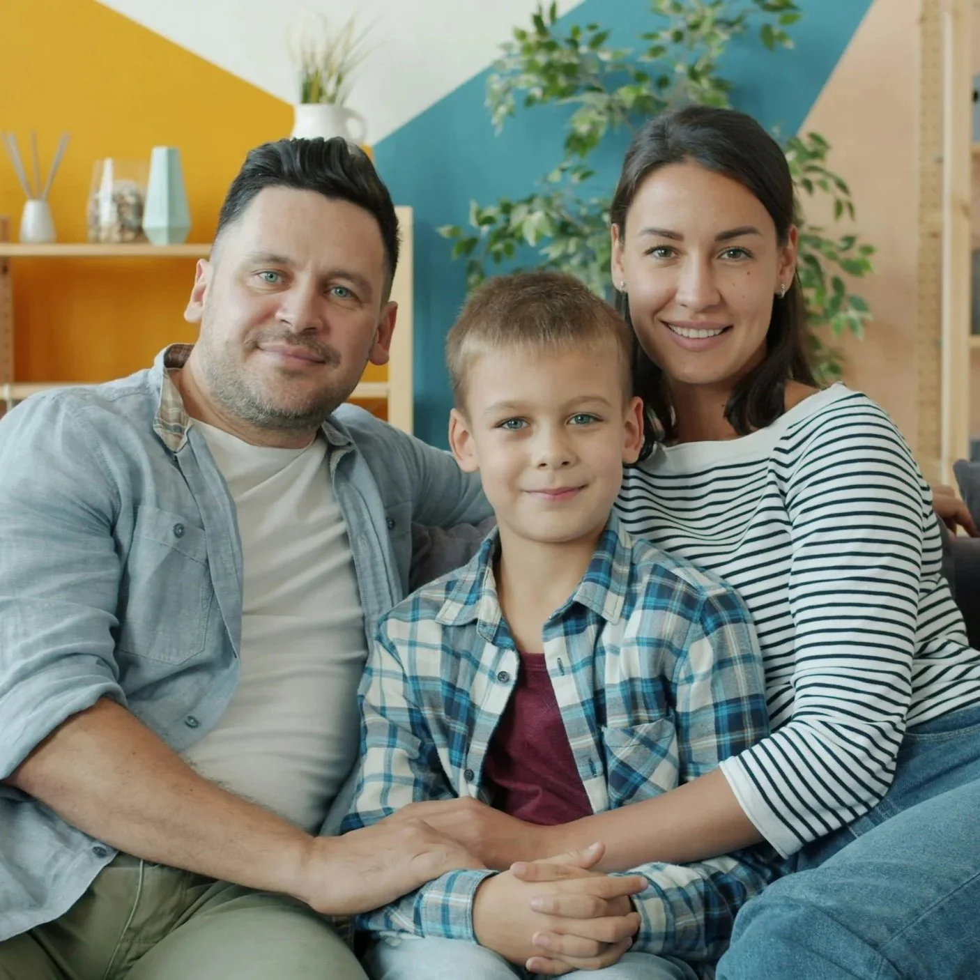 A smiling family of three sitting closely together indoors with a colorful background of teal and yellow walls and a shelf. The family includes a man, woman, and young boy, all looking at the camera.