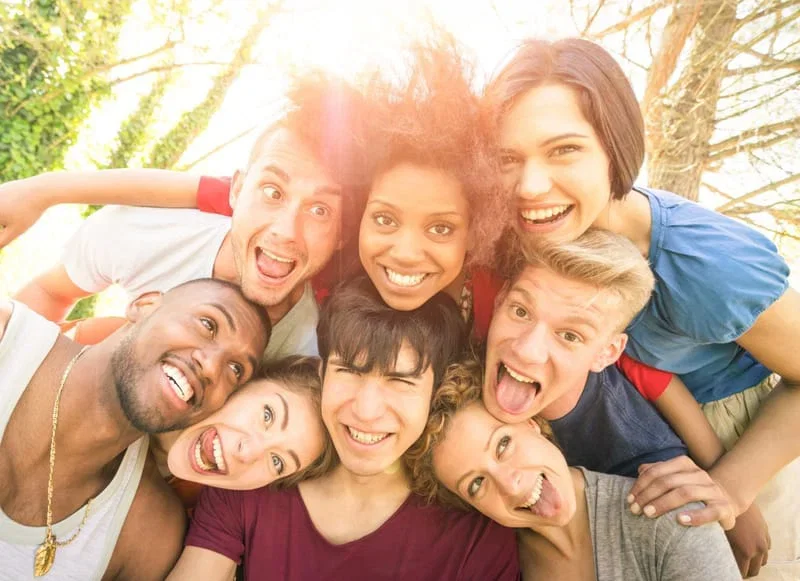 Group of nine diverse young adults smiling and making silly faces outdoors in sunny weather.