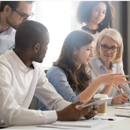 Group of five diverse people in a professional setting having a meeting, with one woman gesturing as she speaks.