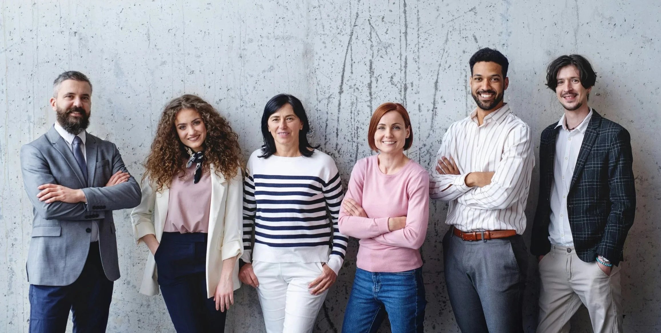 A diverse group of seven people standing against a concrete wall, smiling, with arms crossed or at their sides, dressed in business casual attire.