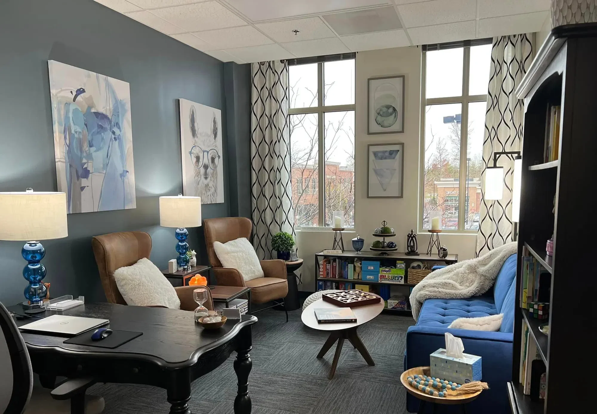 A cozy living room with large windows, decorated with blue, beige, and gray tones, featuring artwork, lamps, a bookshelf, and seating including a blue sofa and brown chairs.