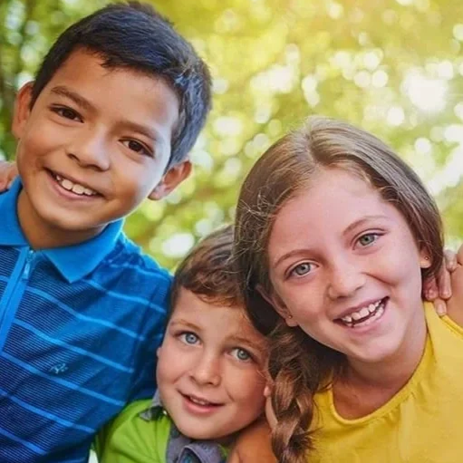 Three kids smiling outdoors with blurred trees in the background.